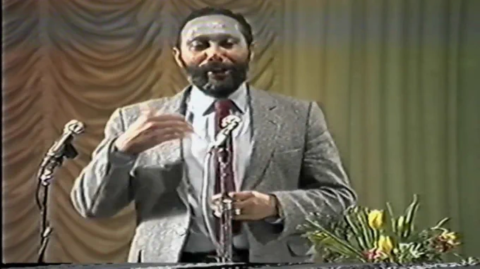 Still from video footage of Professor Stuart Hall giving his 1983 Marx Memorial Lecture. Professor Hall is wearing a grey suit jacket, standing in front of two microphones on stands. To his left (right of the photograph) there are some flowers. The background is a beige curtained wall.