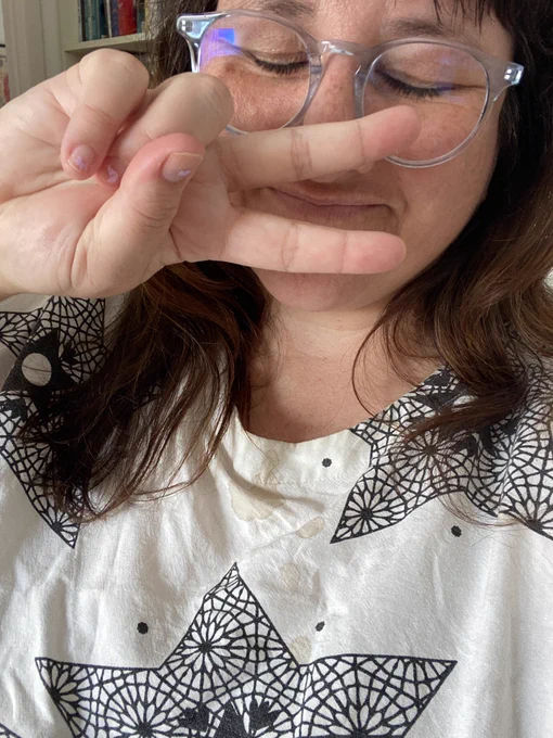 A selfie of me, a white lady with long brown hair and blue glasses on. I’ve got my eyes closed and am giving a peace sign. My white and black dress has three large coffee stains on the front