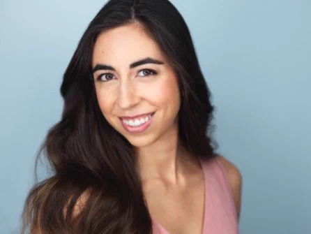 A professional headshot of Eugenia Forteza.  The background is a soft blue.  Forteza wears a smile and a light pink top.  Her long, dark hair covers her right shoulder.  She has tan skin and brown eyes.