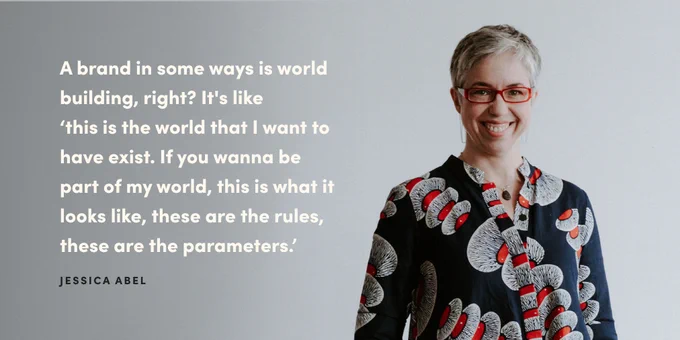 A landscape graphic with a grey background and a portrait photo of Jessica Abel from the mid-torso up on the right hand side. Jessica has short grey hair and is wearing a long-sleeved navy dress that has white and red patterns on it, and red glasses, and she's smiling at the camera. On the left hand side of the graphic, a quote in bold, white, sans serif text reads "a brand in some ways is world building, right? It's like 'this is the world that I want to have exist. If you wanna be part of my world, this is what it looks like, these are the rules, these are the parameters.'" Underneath in black it says Jessica Abel in all caps.