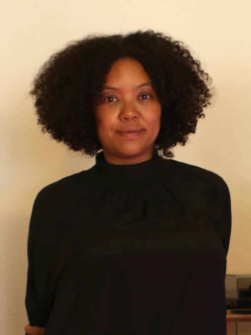  Naomi Velaphi stands in front of a wall and smiles into the camera. She wears a black long-sleeved shirt.