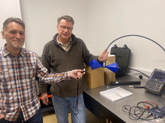 Gareth is holding a vehicle immobiliser key fob and pointing it towards an antenna formed from interesting back to back geometrical shapes, held by Stepan. They are in an electronics lab.