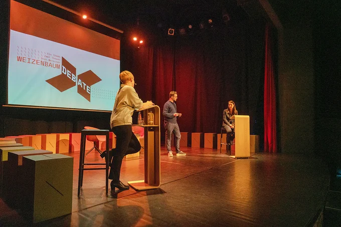 Elisa Lindinger (links), Sascha Friesike (mittig) und Sabine Frank auf der Bühne, im Hintergrund das Logo der Weizenbaum Debate. 