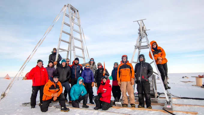 MELT and Icefin teams pose together at their fieldwork site - a white, flat expanse of snowy ice. They are warmly dressed for the cold in orange and red thermal jackets. 16 people are in the photo, two are on ladders.