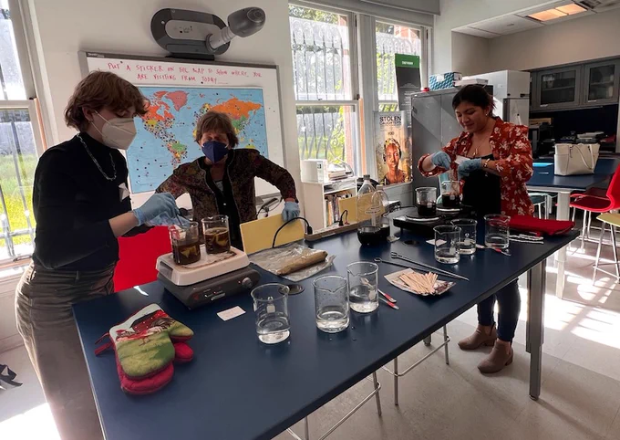 Two participants instructed by the workshop leader at a blue lab bench. They are wearing blue latex gloves while tending to beakers full of dye, stirring a brown liquid. 