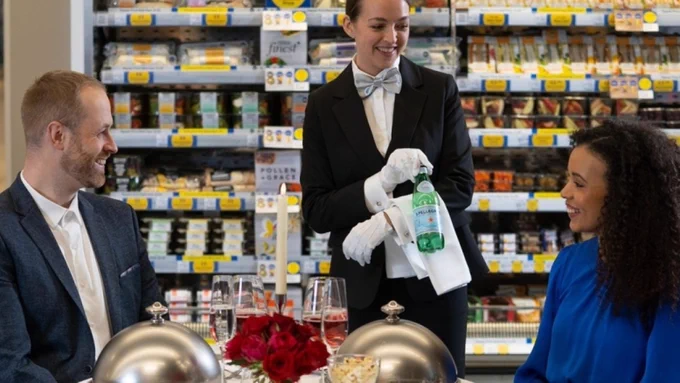 A waitress is presenting a bottle of water to the customers at a table in a supermarket. Copyright: Tesco
