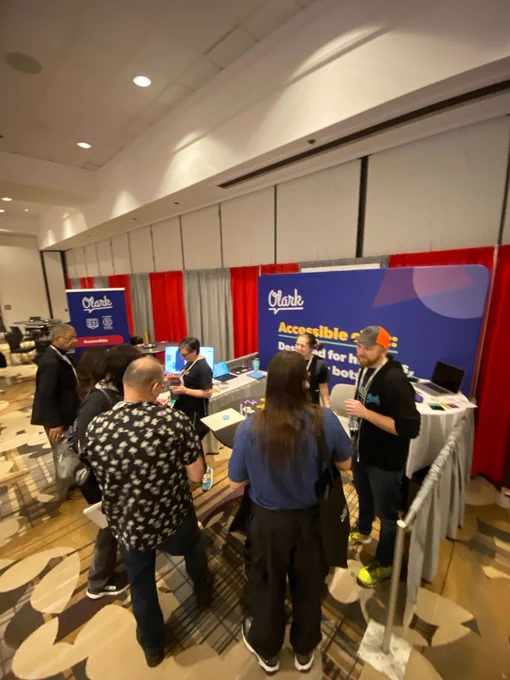 Image: A group of people standing at the Olark booth #1008, at CSUN 2023. They are smiling and engaging with each other, some are looking at a computer screen displaying Olark's platform, and others are speaking with Olark reps. The booth features a sign that reads 'Olark Accessible Chat'.
