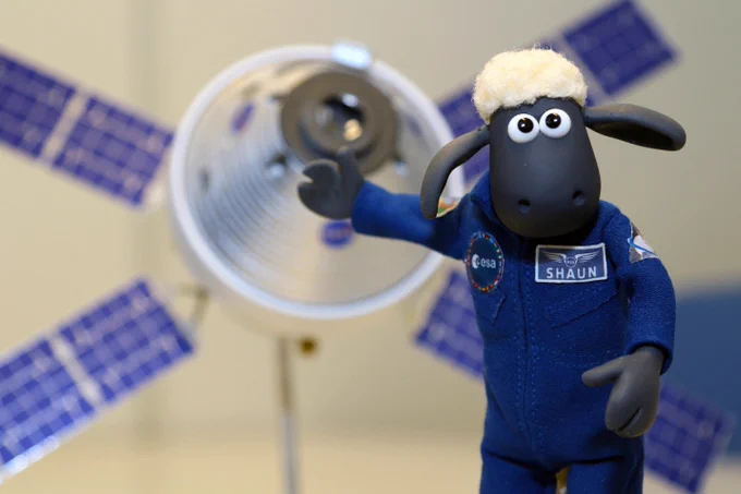A small clay sheep wearing a blue astronaut uniform stands in front of an image of a satellite.
