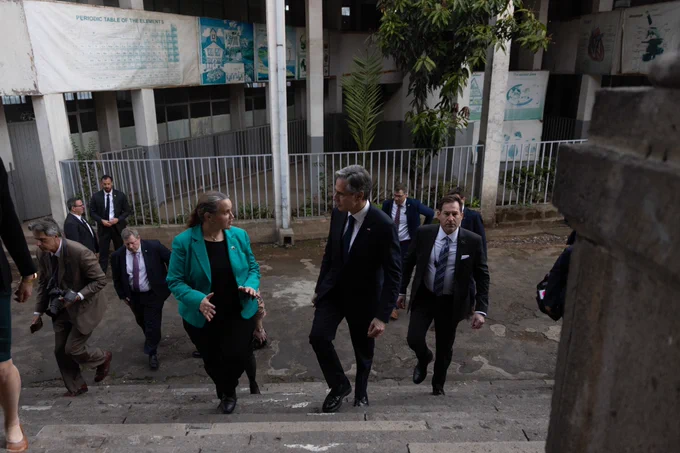 Secretary Blinken speaks to a woman wearing a green blazer as they walk up a stairs to enter a building. A group of advisors, security, and meeting participants follows them.