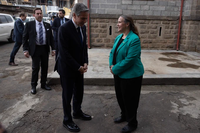 Secretary Blinken speaks to a woman wearing a green blazer as they stand outside a large building.