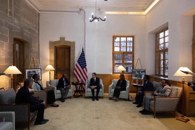 Secretary Blinken sits and talks with a group of human rights leaders and advocates in upholstered chairs arranged in a half circle configuration. An American flag sits behind them.