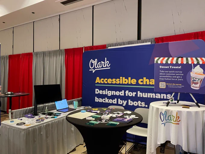 A picture of the Olark accessible chat booth #1008 at CSUN conference. The booth is adorned with a banner that reads 'Olark accessible chat designed for humans, backed by bots', and features free swag on the tables, such as stickers, and buttons. There is a mini poster offering a free treat via a survey.