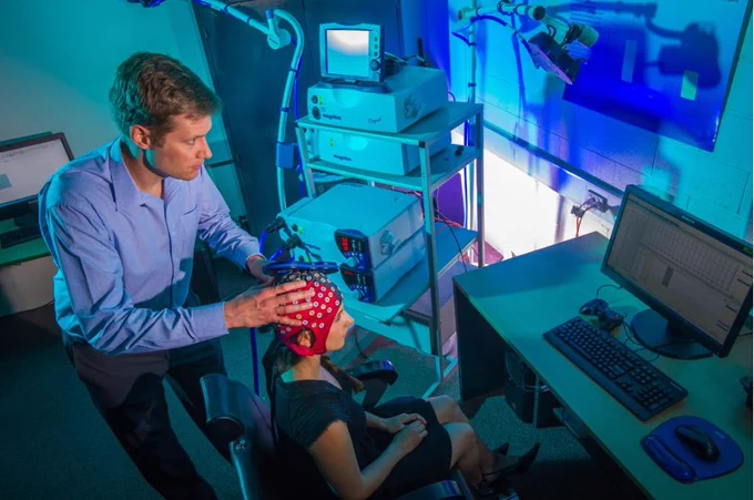 Two people in a room with the TMS machine. One person is standing behind the other person who is sitting in a chair facing a computer. The person standing is delivering TMS intervention to the person sitting