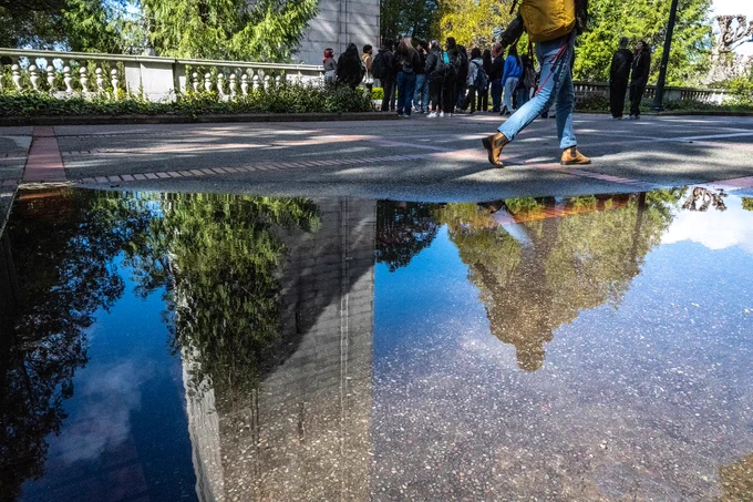 Photo of a puddle with the water reflecting the Campanile on the campus of the University of California, Berkeley. People are seen looking at the Campanile and walking by the puddle.