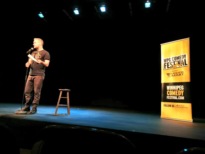 Dean Jenkinson at a microphone on the stage of Gas Station Arts Centre in front of a Winnipeg Comedy Festival Yellow Banner 