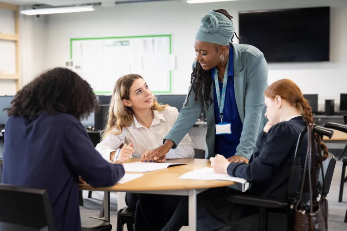 Three students sitting together in the classroom. One of the students is speaking with the teacher who is standing next to a table.