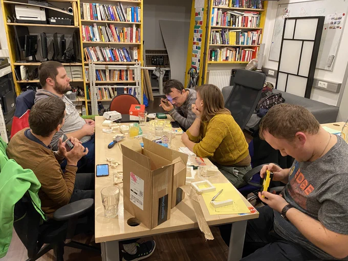 Sechs Menschen sitzen an einem Tisch. Sie beschäftigen sich mit Tastaturen und Smartphones in ihren Händen oder verfolgen die Arbeit der Person nebenan. Auf dem Tisch stehen Trinkgläser, geöffnete Kartons, Smartphones, Tastaturen, Klemmbausteine, Knetmasse und noch mehr Arbeitsmaterial. In den Regalen im Hintergrund stehen geschlichtete Bildschirme, Tastaturen, Drucker und viele Bücher. An der rechten Wand steht ein Sofa, eine Wandteiler und ein Whiteboard direkt auf der Wand.