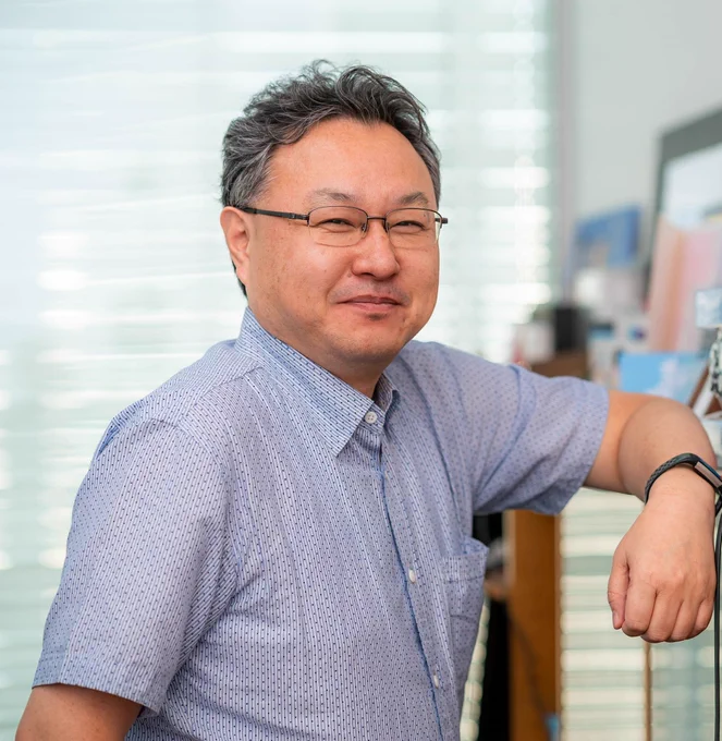 Shuhei Yoshida poses for a photograph. He wears a blue, short sleeved shirt and glasses whilst leaning against an offscreen cabinet. He smiles warmly at the photographer. 