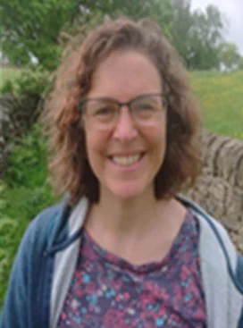 Laura smiles at the camera. They have shoulder length gurly brown hair, is wearing glasses and a pink and blue top.