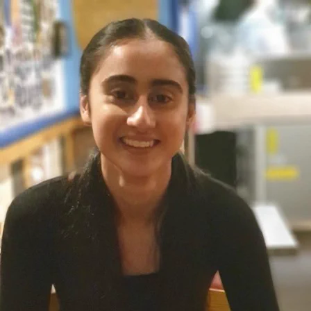 Simran smiles at the camera. They have long dark hair and is wearing a black top.