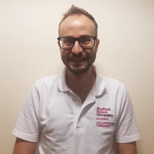 Oli smiles at the camera. They are wearing glasses, has brown hair and is wearing a white shirt.