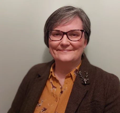 Natalie smiles at the camera. They have short dark hair, is wearing glasses and a brown top.