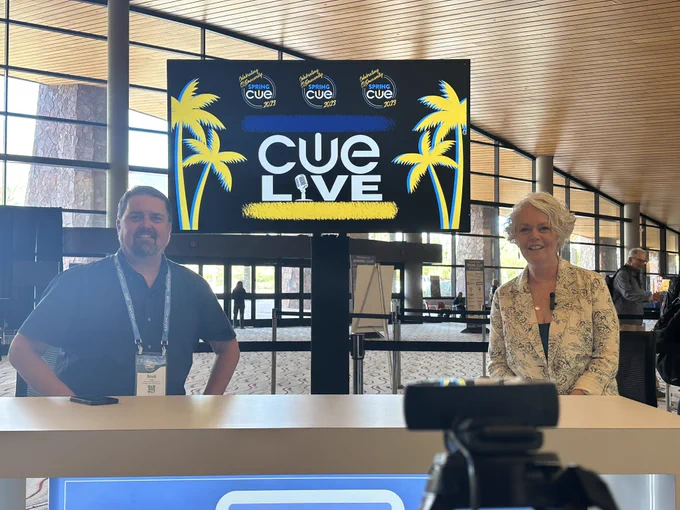 Man and woman seated at a table with CUE LIVE on a screen behind them, in a convention center.
