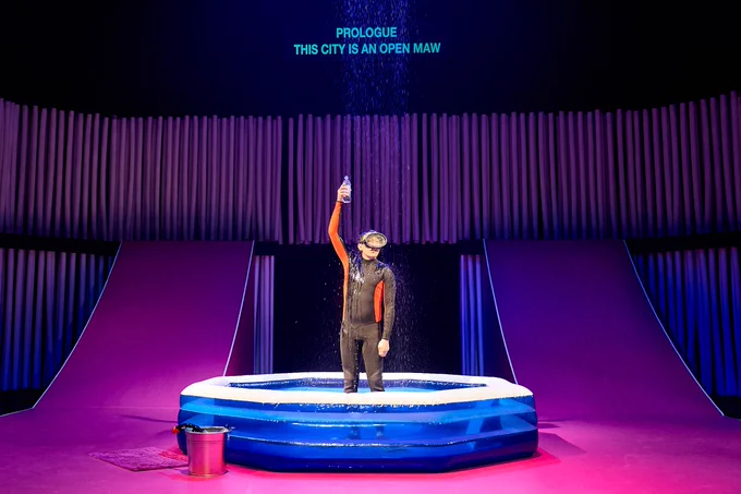 Leo Wan in The Good Person of Szechwan. Photo by Manuel Harlan. Leo wears a wetsuit and stands in a paddling pool as water rains down over them. They hold up a plastic bottle to capture some of the water.