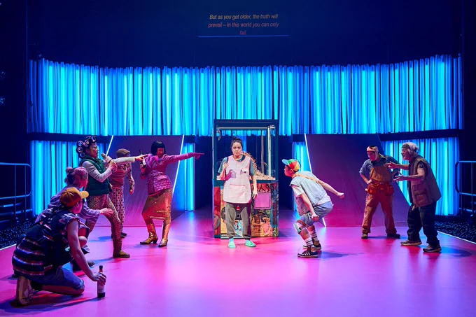 Ami Tredrea and the Company in The Good Person of Szechwan. Photo by Manuel Harlan. Ami stands in front of a claw grabber machine filled with cigarettes. They wear casual clothes and behind them is lit up in blue light. Around them are cast members in bright clothing, dancing and waving their arms.