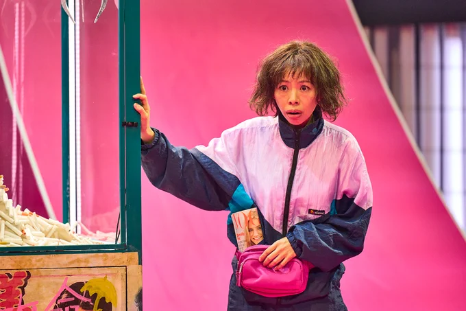 Suni La in The Good Person of Szechwan. Photo by Manuel Harlan. Suni wears a sports jacket and leans against a claw grabber machine filled with cigarettes. They look perplexed.