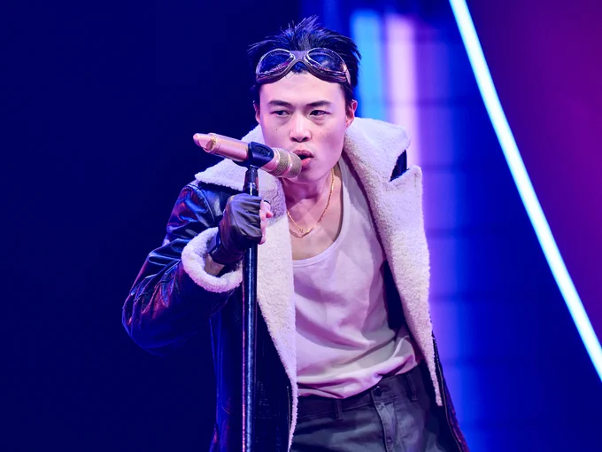 Aidan Cheng in The Good Person of Szechwan. Photo by Manuel Harlan. Aidan wears an aviator jacket and goggles and sings into a gold microphone on a stand.
