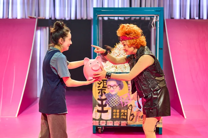 Ami Tredrea and Melody Brown in The Good Person of Szechwan. Photo by Manuel Harlan. Melody wears leather studded biker clothes and hands a pink piggy bank to Ami, who takes it with a smile. They are in front of a claw grabber machine filled with cigarettes.