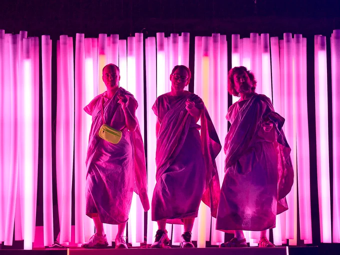 Tim Samuels, Nick Blakeley and Callum Coates in The Good Person of Szechwan. Photo by Manuel Harlan. Against a neon pink background, three gods dressed in white togas perform a synchronised dance routine.