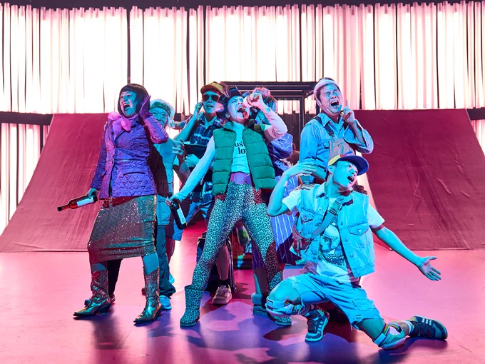 The Company of The Good Person of Szechwan. Photo by Manuel Harlan. A group of people wearing brightly coloured clothes gather in a group and sing karaoke. They scream into microphones and hold their arms out as they sing and dance.