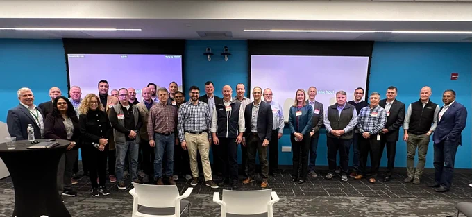 Over two dozen people stand in front of two projector screens and pose for photo. Majority of people are men. 