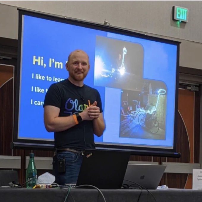 Image: A picture of Olark CEO, Ben Congleton, standing in front of a presentation deck that reads 'Hi, I'm Ben'. He is casually dressed in a black t-shirt adorned with the Olark logo in rainbow colors, paired with comfortable blue jeans, and is smiling with his hands clasped together in a friendly way.