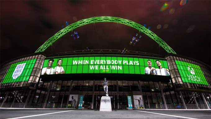The front entrance of Wembley Stadium is lit up green, the center board featuring England and Lionesses players, has text reading "When Everybody Plays, We All Win" with England and Xbox logos either side.