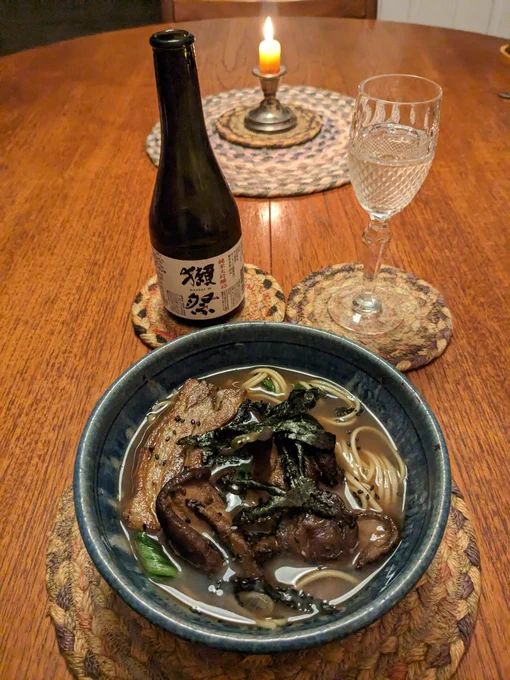 Photo of a dining table. On it stands a bowl of ramen in a brown broth with nori and pork slice. Behind the bowl is a bottle of sake and a glass of sake, behind the bottle is a flickering candle 