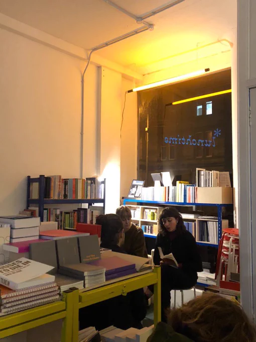 Two people sit in the corner of a bookshop with books all around them