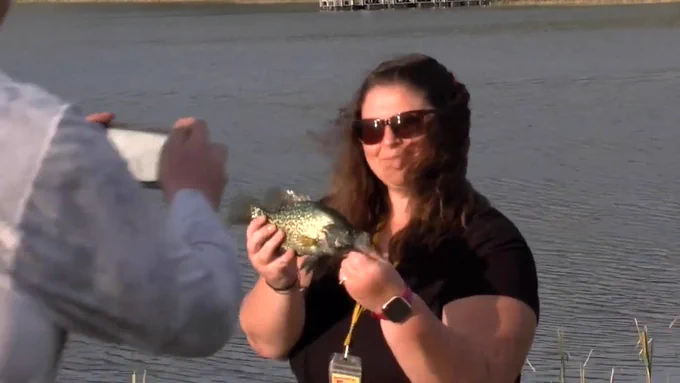 A woman poses for a picture while holding a crappie she caught while fishing. A lake is in the background behind her.