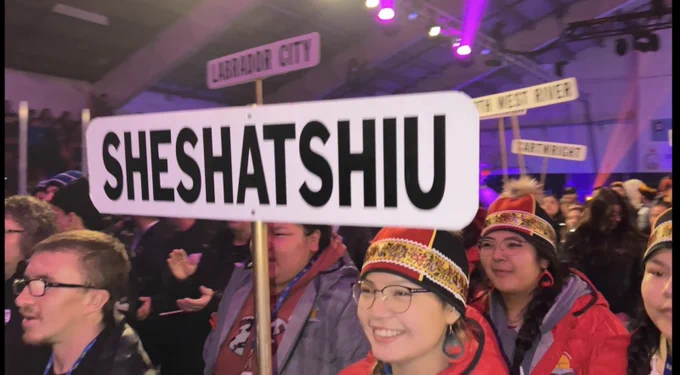 Teams walk through arena carrying community name signs.