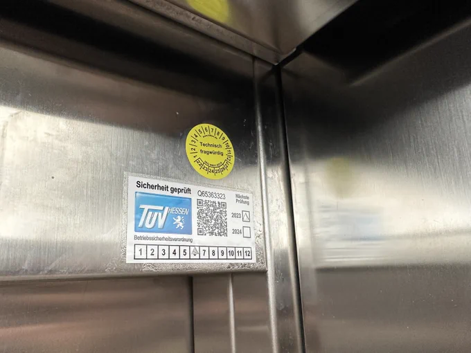 Ein Bild von einem gelben runden sticker, mit „technisch fragwürdig“ in der mitte. In der Ecke eines aufzuges.