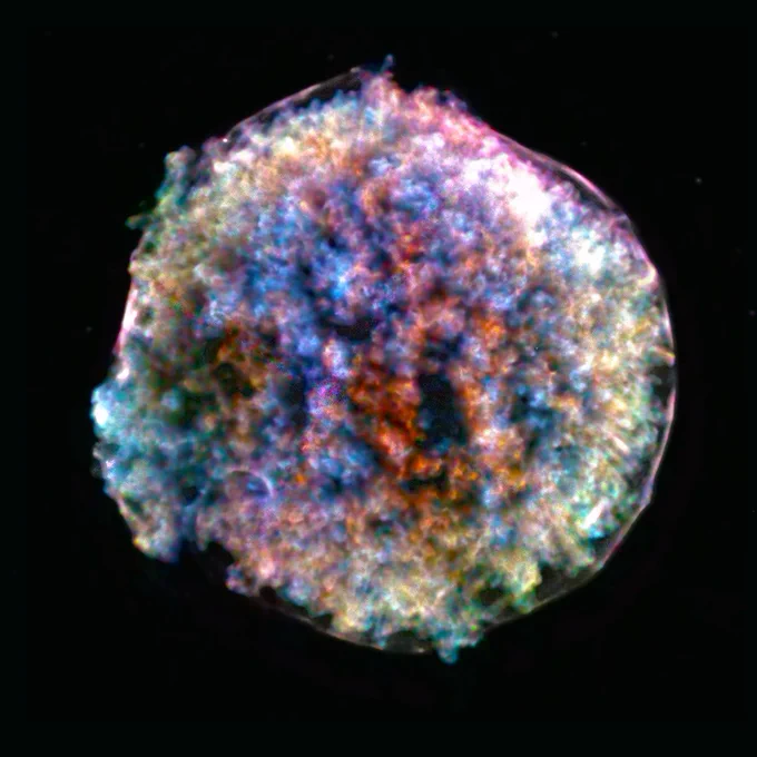This is an X-ray image of Tycho's supernova remnant. The remnant is mostly round in shape, and it resembles a fluffy, clumpy cotton ball set against a black background. Its surface is a mottled mix of reds, pinks, greens, blues, and yellows with tufts of color tightly packed into the fuzzy-looking ball.