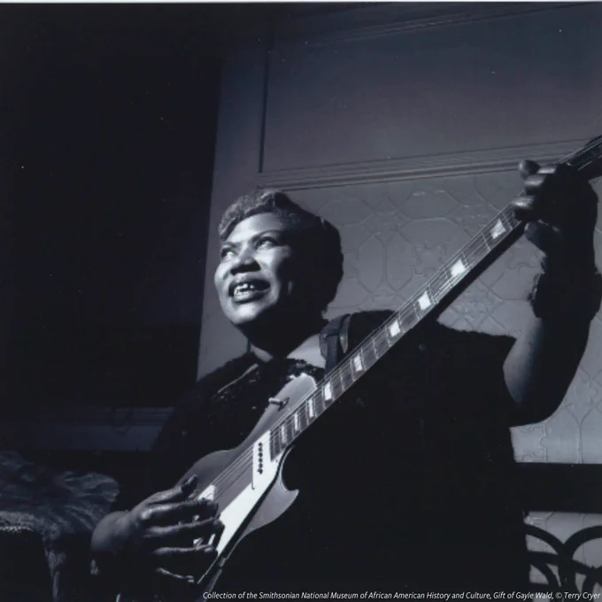 A black-and-white photograph of Sister Rosetta Tharpe. She is wearing a black shirt and sitting. She is holding a guitar and looking up and to her right. A fur coat is on the left side of the image and white, patterned wall and darkened window are in the back ground. 