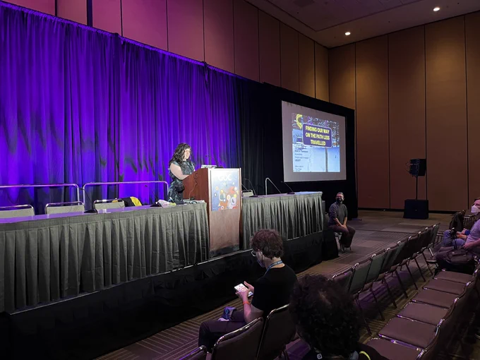 Photo of Aderyn Thompson on stage at GDC. The title slide says “Finding our way on the path less travelled”