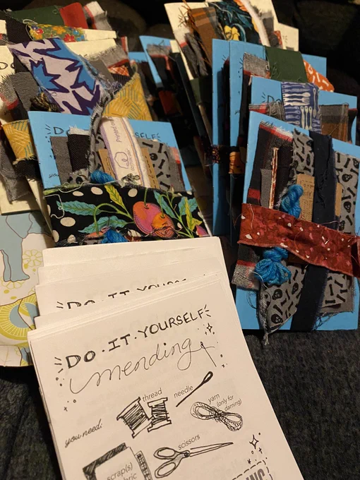 A bunch of mending kits with fabric, yarn, thread, pins, and zines, plus a stack of zines titled Do It Yourself Mending with drawings of needed tools and materials