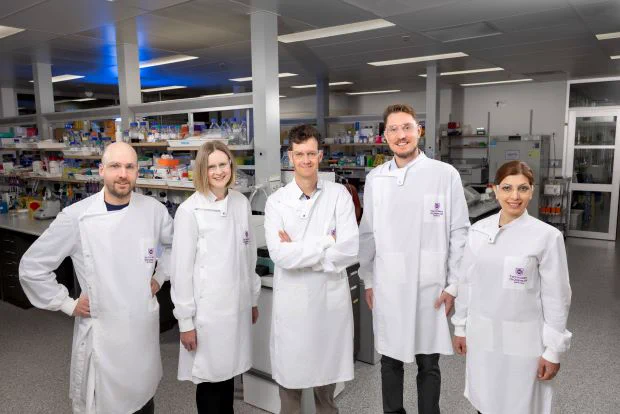 A group of five male and female scientists in white coats stand with a lab in the background.