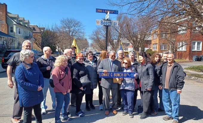 Group of adults at neighborhood intersection, dressed warmly. At center, Lonnie Bunch smiles. He holds a sign that says Lonnie G. Bunch III Way. Behind the group, a similar sign is on sign post.