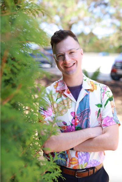 Eric Balboni stands outside with his arms folded and a smile on his face.  He is fair-skinned with dark hair.  He wears glasses with transparent frames, a dark t-shirt under a colorful short-sleeved button down, a brown belt, and dark pants.  Greenery is visible to his right and in the background of the photo.