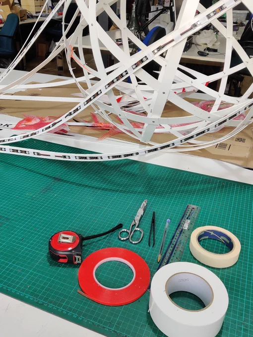 A selection of tapes with scissors, tweezers, a pen, ruler and tape measure, on a green cutting mat in front of a rectified truncated icosahedron made from PVC strips, with extra PVC streamers which have LED strips attached to them